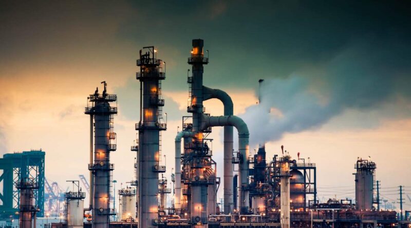 A steaming, glowing mass of smoke stacks and distillation towers connected by conduit and pipe racks towers in the early morning light in front of the Port of Los Angeles.