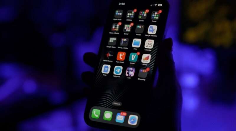 a photo in a darkened room of an iPhone screen lit up in dark mode with a bunch of apps on the screen.