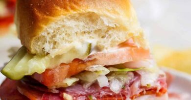 A small sandwich with sliced meats, cheese, tomato, lettuce, pickles, and mayonnaise on a soft bun, shown with a bite taken out of it.