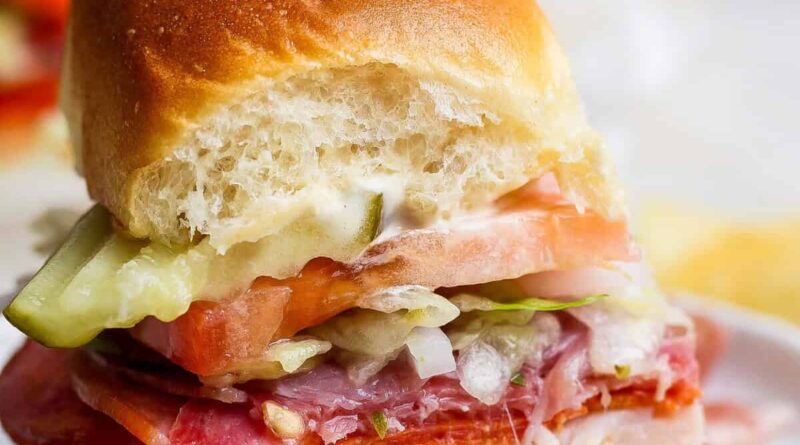 A small sandwich with sliced meats, cheese, tomato, lettuce, pickles, and mayonnaise on a soft bun, shown with a bite taken out of it.