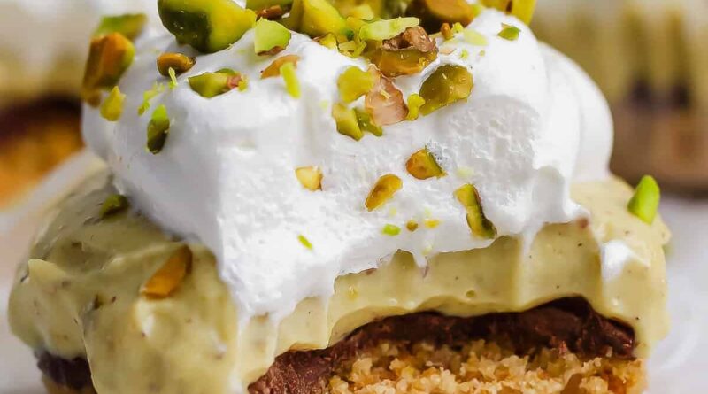 A dessert bar with a crumbly base, chocolate layer, creamy filling, whipped topping, and chopped pistachios, with a bite taken out of it.
