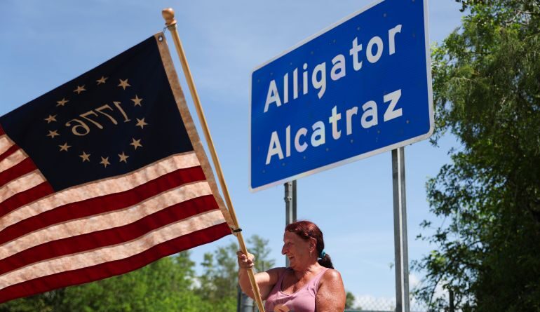 A person waves a US flag in front of a sign for Alligator Alcatraz.