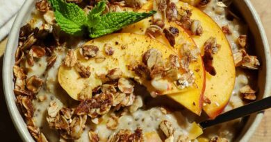 A bowl of oatmeal topped with granola, peach slices, and a sprig of mint, with a spoon placed inside.