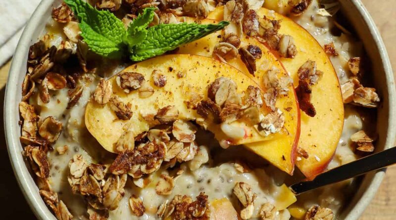 A bowl of oatmeal topped with granola, peach slices, and a sprig of mint, with a spoon placed inside.