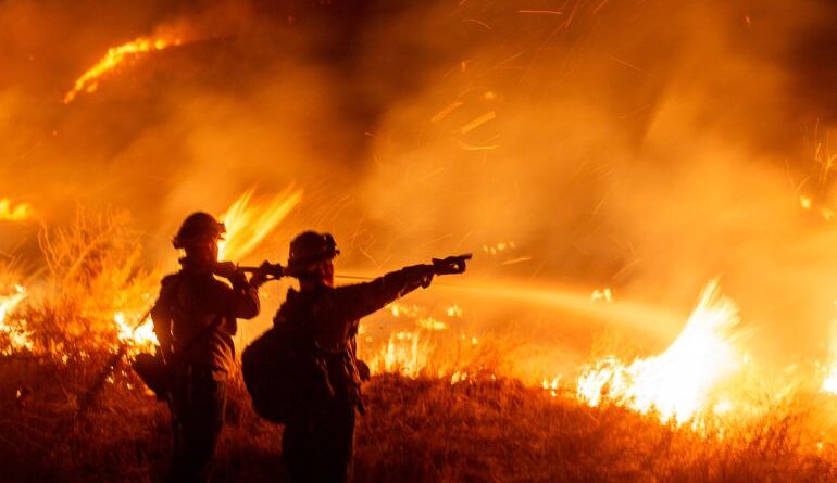 A pair of firefighters stand silhouetted against a burning blaze at night: One points.