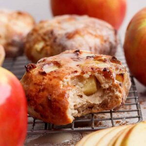 Close-up of a glazed apple pastry, reminiscent of air fryer apple fritters, with a bite taken out, resting on a cooling rack and surrounded by fresh apples and apple slices.