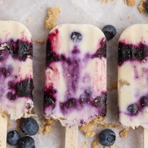 Three creamy blueberry popsicles with purple swirls are arranged on a light surface, surrounded by scattered blueberries and crumbled cookie pieces.