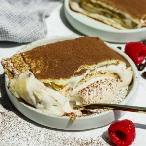 A plate of cottage cheese tiramisu crepe roll with a spoonful taken out, topped with cocoa powder, sits next to fresh raspberries on a white surface.