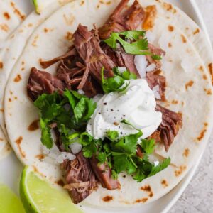 A flour tortilla topped with shredded beef, chopped onions, fresh cilantro, and a dollop of sour cream, served with a lime wedge.