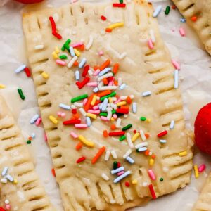 A rectangular pastry topped with icing and colorful sprinkles sits on parchment paper next to a fresh strawberry.