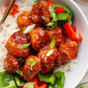 A bowl of white rice topped with crispy glazed chicken pieces, sliced green onions, and red and green bell peppers.