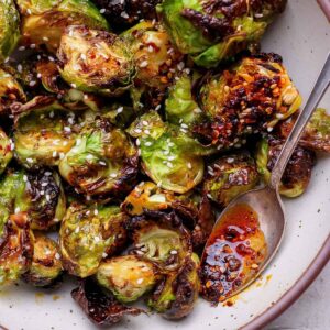 Roasted Brussels sprouts with a glossy chili sauce, garnished with sesame seeds, served in a bowl with a spoon.