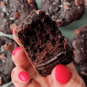 A hand with pink nail polish holds a bitten Kodiak protein muffin, revealing its moist, dense interior; more chocolate muffins are blurred in the background.