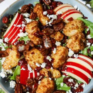 A salad with mixed greens, sliced red apples, breaded chicken pieces, pecans, dried cranberries, crumbled feta cheese, and a light drizzle of dressing.