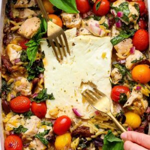 A baked dish with feta cheese in the center, surrounded by cherry tomatoes, cooked chicken pieces, orzo, spinach, red onions, olives, and fresh basil, with two forks serving.