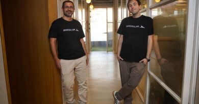 The two co-founders stand abreast in an office hallway wearing black logo t-shirts.
