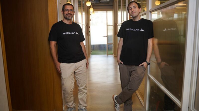 The two co-founders stand abreast in an office hallway wearing black logo t-shirts.