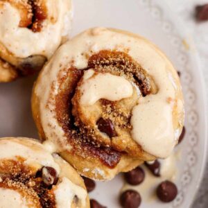 Close-up of cinnamon rolls topped with icing and sprinkled with brown sugar, with chocolate chips scattered on a white plate.