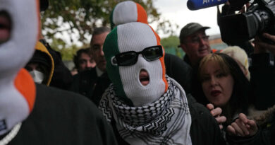 Liam Óg Ó hAnnaidh wears sunglasses, a balaclava in the colors of the Irish flag, and a kaffiyeh as a scarf while walking through a crowd of people.