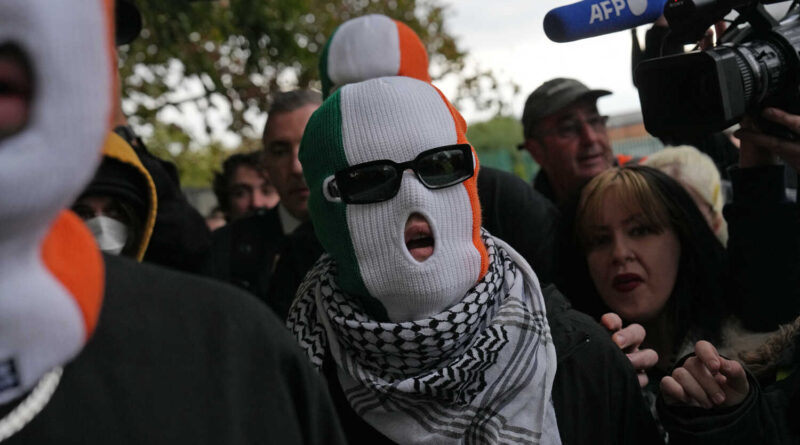 Liam Óg Ó hAnnaidh wears sunglasses, a balaclava in the colors of the Irish flag, and a kaffiyeh as a scarf while walking through a crowd of people.