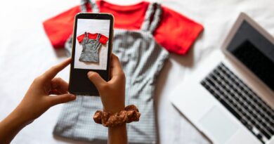 A girl uses her phone to take a photo of two items of clothing, a red tee-shirt and striped dungarees, to sell online on a nearby laptop