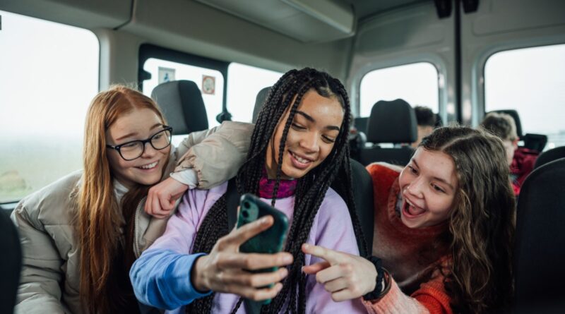 A front view of a group of three teenage girls on the minibus on the way to go on a hiking field trip. They are looking at one of the girls mobile phones and smiling and pointing at the screen.