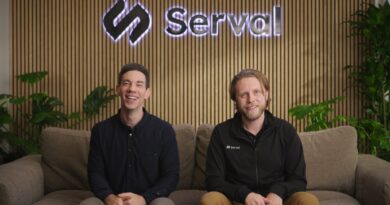 Two caucasian men sit on a couch with a company logo behind them.