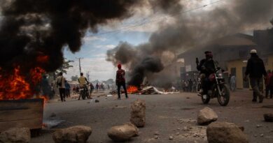 Protesters stand and sit on motorbikes as smoke billows from two fires burning on the street.