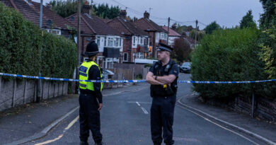Two police officers stand in the middle of a road blocked by police tape, with houses in the background and shrubs lining the sidewalks.
