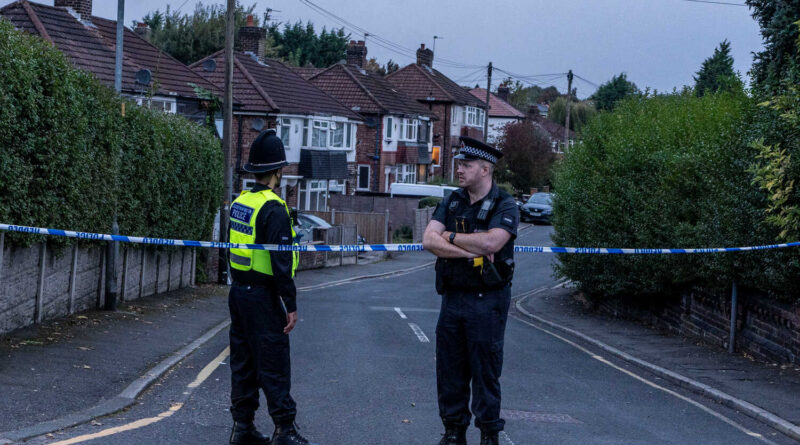 Two police officers stand in the middle of a road blocked by police tape, with houses in the background and shrubs lining the sidewalks.