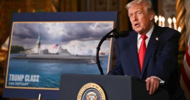 Donald Trump speaks at a podium beside a display of the “Trump Class” USS Defiant warship concept.