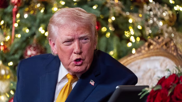 Donald Trump speaks with a serious expression, seated near a decorated Christmas tree and holiday lights.
