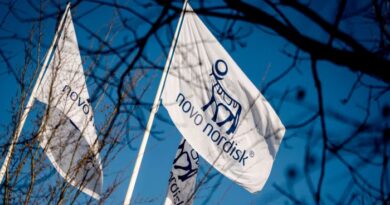 Flags displaying the Novo Nordisk logo and name fly against a blue sky, partially obscured by tree branches.