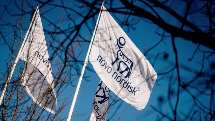 Flags displaying the Novo Nordisk logo and name fly against a blue sky, partially obscured by tree branches.