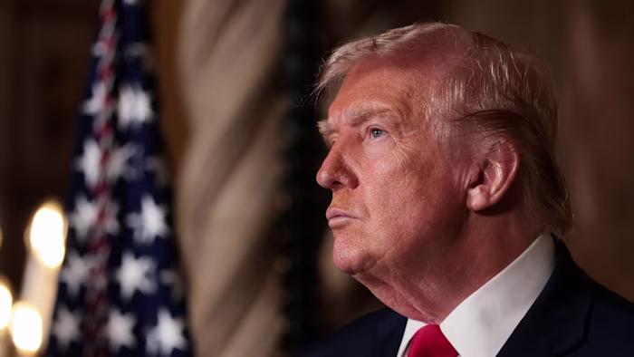 Donald Trump appears in profile, looking serious, with a US flag blurred in the background.