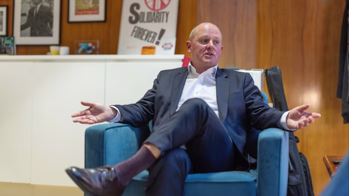 Paul Nowak sits in a chair with arms outstretched during an interview at the TUC head office, with union posters visible behind him.