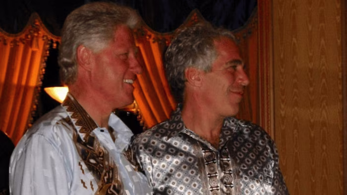 Bill Clinton stands beside Jeffrey Epstein, both smiling and wearing patterned shirts, in an indoor setting with gold drapes behind them. (Image supplied by the DoJ)