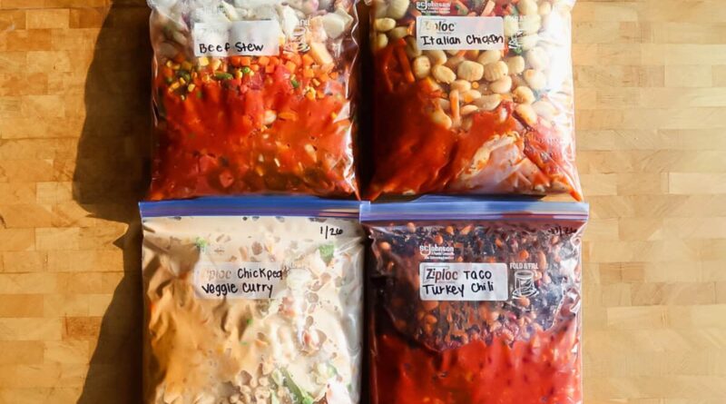 Four labeled freezer bags filled with homemade meals&mdash;beef stew, Italian chicken, chickpea veggie curry, and taco turkey chili&mdash;arranged on a wooden surface.