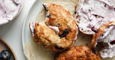 A blueberry bagel with a bite taken out, filled with lavender-colored cream cheese, rests on parchment paper beside another bagel and a small bowl of blueberries.