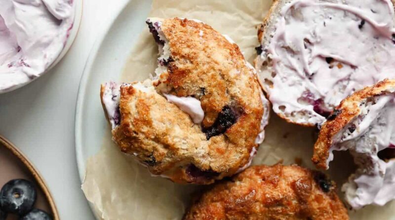 A blueberry bagel with a bite taken out, filled with lavender-colored cream cheese, rests on parchment paper beside another bagel and a small bowl of blueberries.