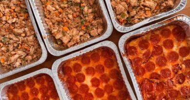 Six aluminum trays, three filled with pepperoni pizza and three with a stir fry dish of vegetables and chicken, are arranged on a wooden surface&mdash;perfect alongside comforting casseroles for any gathering.