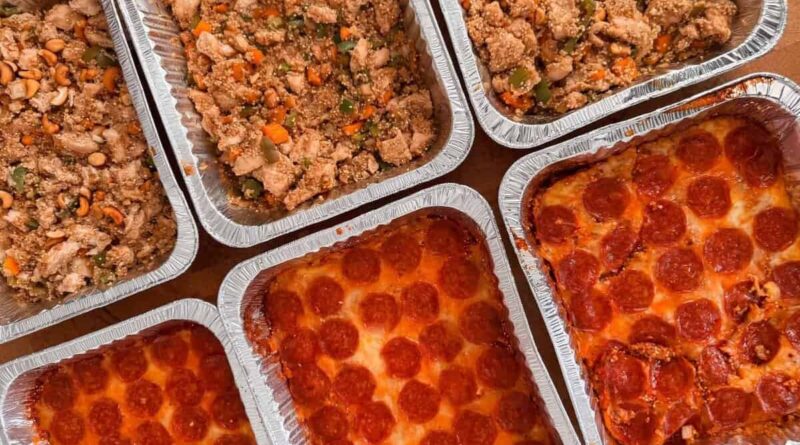 Six aluminum trays, three filled with pepperoni pizza and three with a stir fry dish of vegetables and chicken, are arranged on a wooden surface&mdash;perfect alongside comforting casseroles for any gathering.