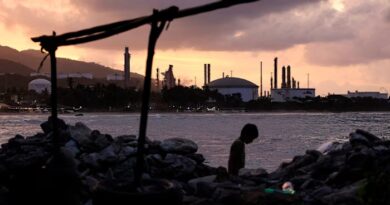 A child walks along a rocky beach at dusk, with the El Palito refinery's industrial structures silhouetted in the background.