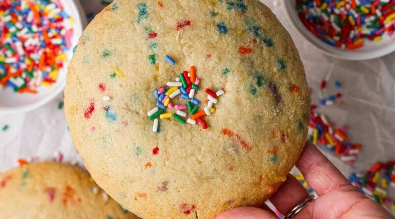 A hand holds a large sugar cookie with colorful sprinkles, with bowls of additional sprinkles and another cookie in the background.