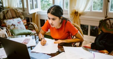 hispanic latina college student works on assignment at home. She is writing something on paper. A laptop is on her desk.