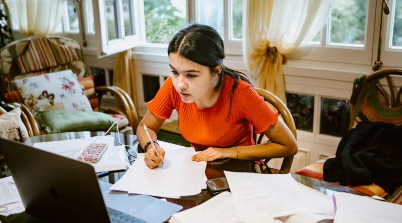 hispanic latina college student works on assignment at home. She is writing something on paper. A laptop is on her desk.