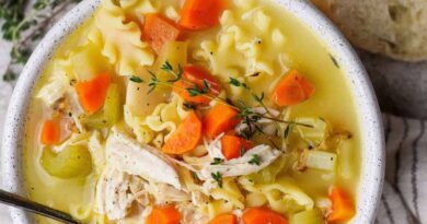 A bowl of chicken noodle soup with carrots, celery, noodles, shredded chicken, and fresh thyme, served with a slice of bread.
