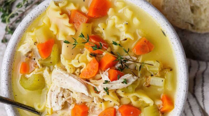 A bowl of chicken noodle soup with carrots, celery, noodles, shredded chicken, and fresh thyme, served with a slice of bread.