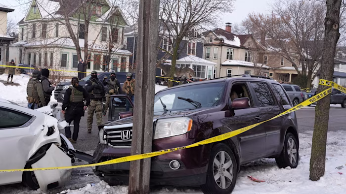 Federal agents in tactical gear stand near a maroon SUV with a bullet hole in the windshield and a damaged white car, behind yellow police tape.