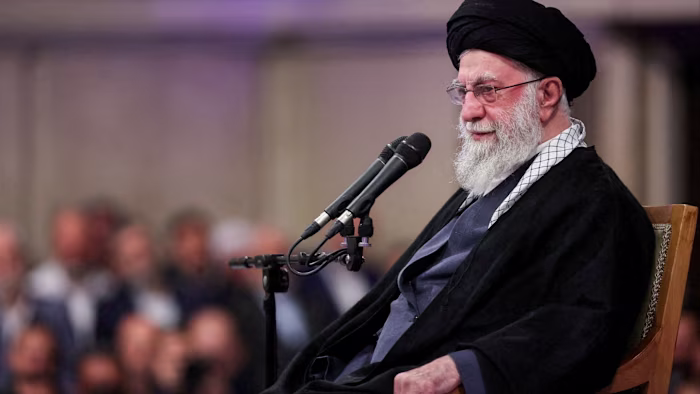 Ayatollah Ali Khamenei speaks into two microphones while seated, with a blurred audience in the background.